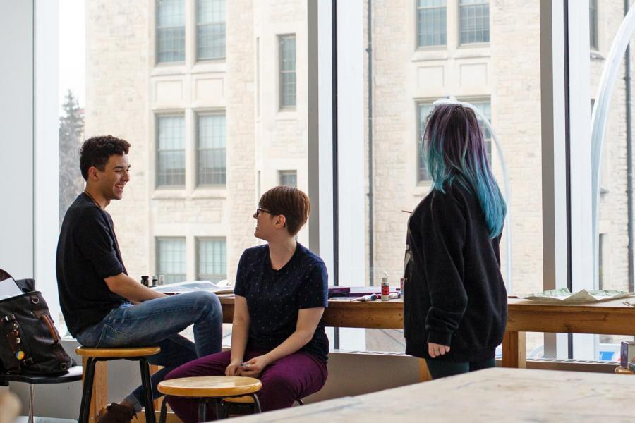 Three School of Art students sit at a narrow table in front of a window. 
