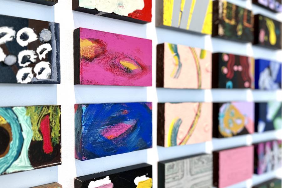 A series of colourful 5x3" painted blocks hang in a grid formation on a white wall.