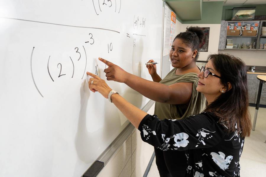 A teacher and student work together to solve a math problem on a whiteboard in a classroom. 