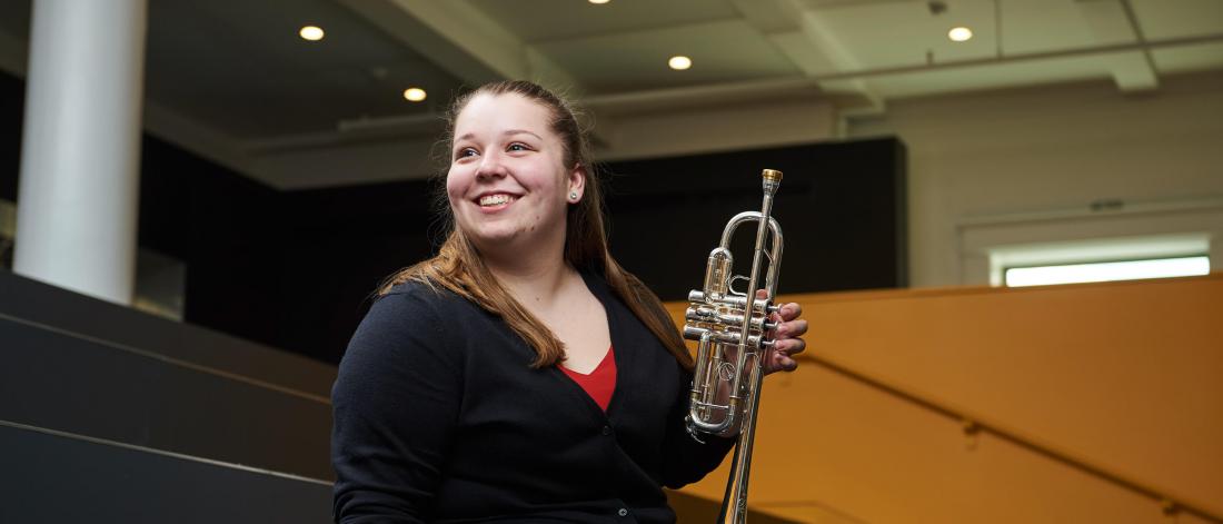 A smiling student sits in the Music library concourse holding a trumpet.