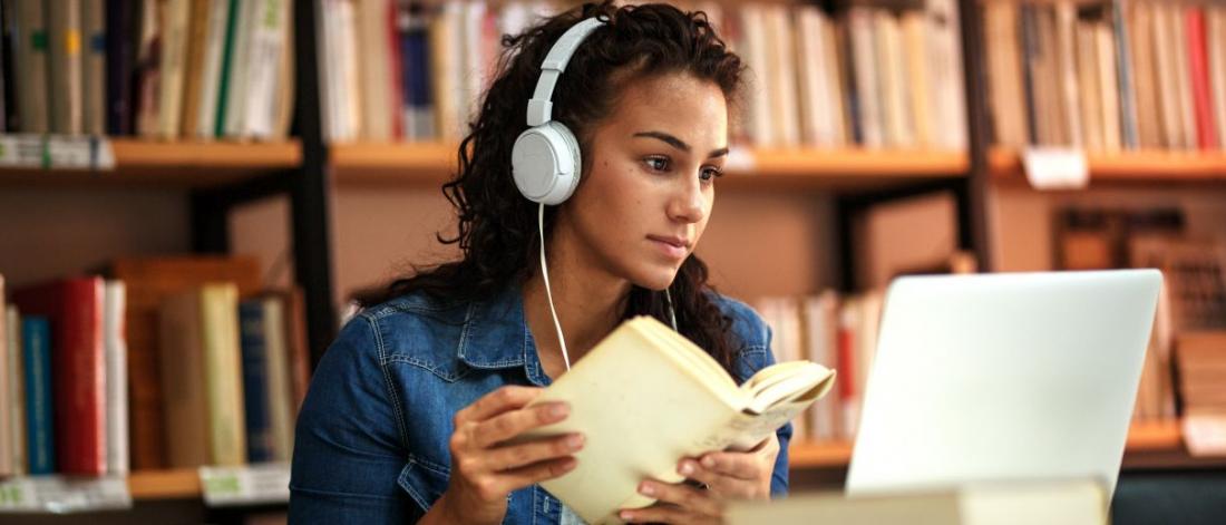 Student looking at laptop wearing white headphones and holding an open book