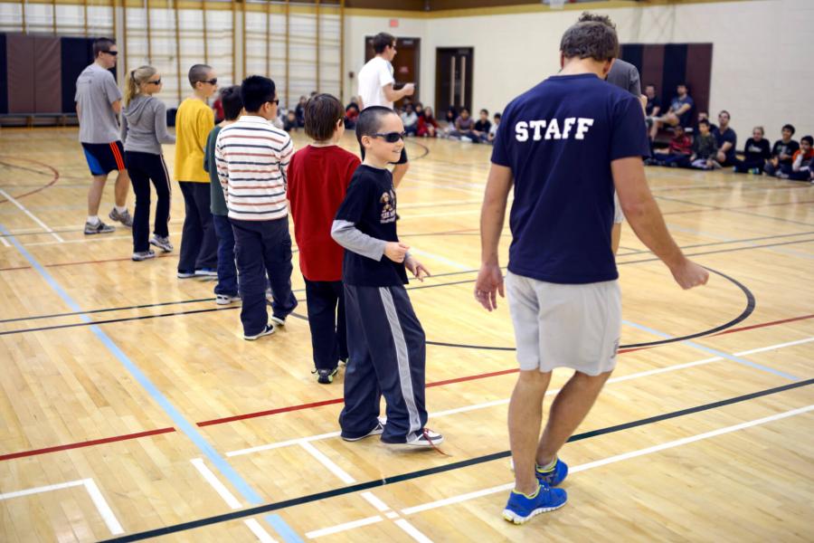 A physical education student teacher demonstrates a skill to a class of young children standing in a row in a gymnasium.