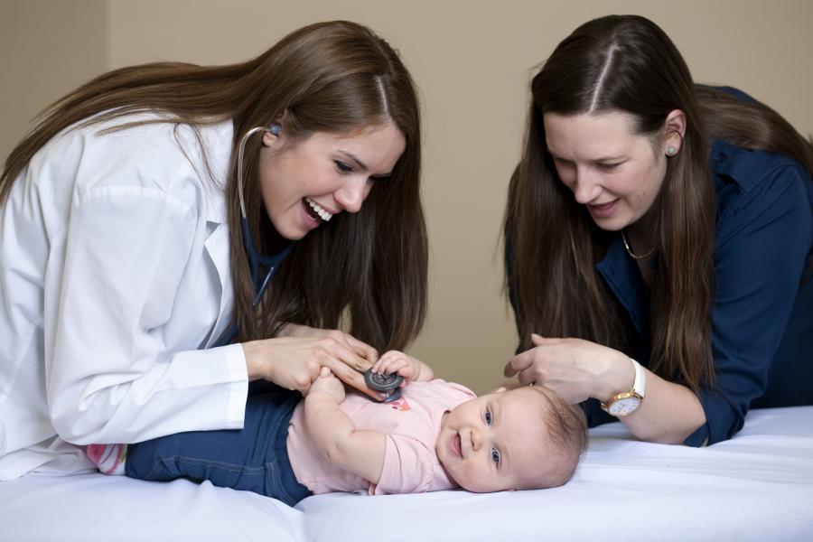 Two students examine a smiling baby girl laying on a table. Both students are smiling as one of them listens to the baby's heartbeat with a stethoscope.