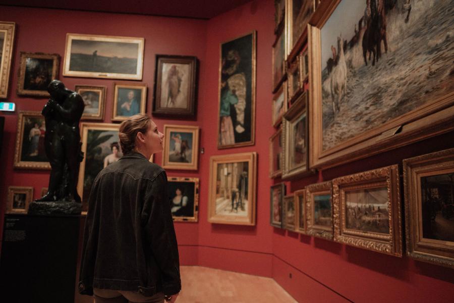 Photo of woman looking at old paintings on a deep red gallery wall.