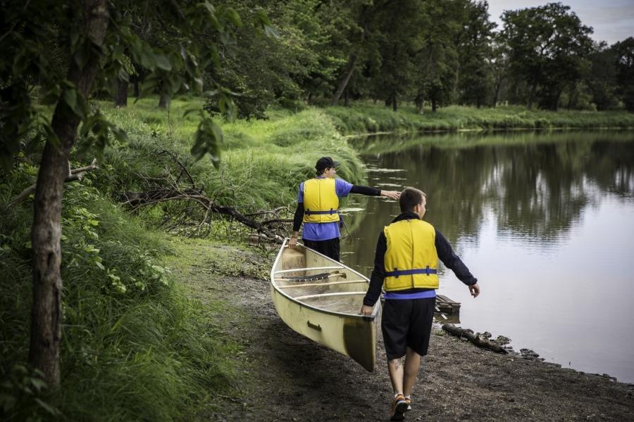 Two men wearing life jackets carrying a canoe at a lakeshore.