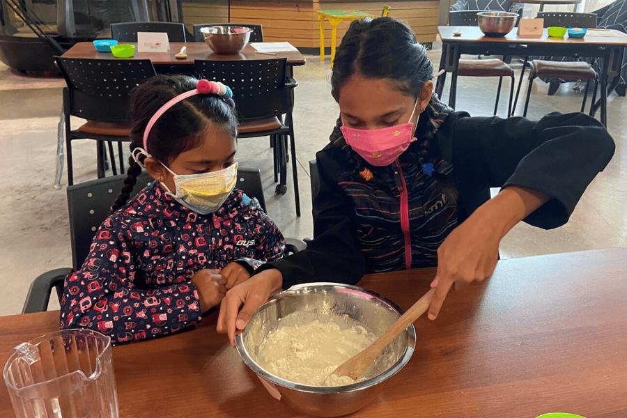 2 children sit at a wooden table wearing masks. One is mixing pizza dough in a metal bowl with a wooden spoon while the other watches.
