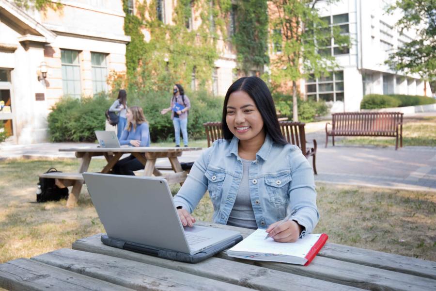A student takes notes while studying at her laptop outdoors seated at a picnic table. 