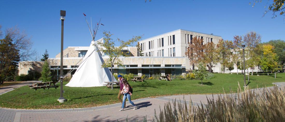 A teepee stands outside of Migizii Agamik - Bald Eagle Lodge during summer months while students walk by and relax at picnic tables.