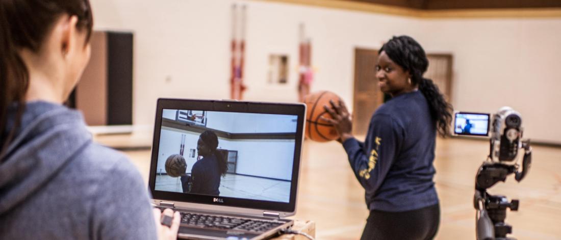 Two researchers in a gymnasium, one stands in front of a laptop and a camera on a tripod while the other prepares to throw a basketball.