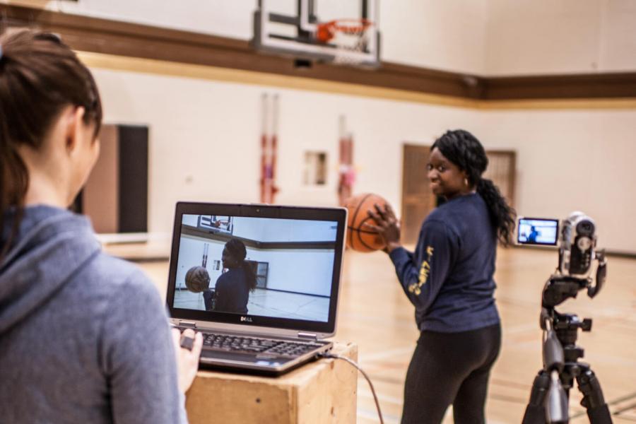 Two students in a gymnasium, one holds a basketball and prepares to throw the ball while the other monitors a laptop and camera.