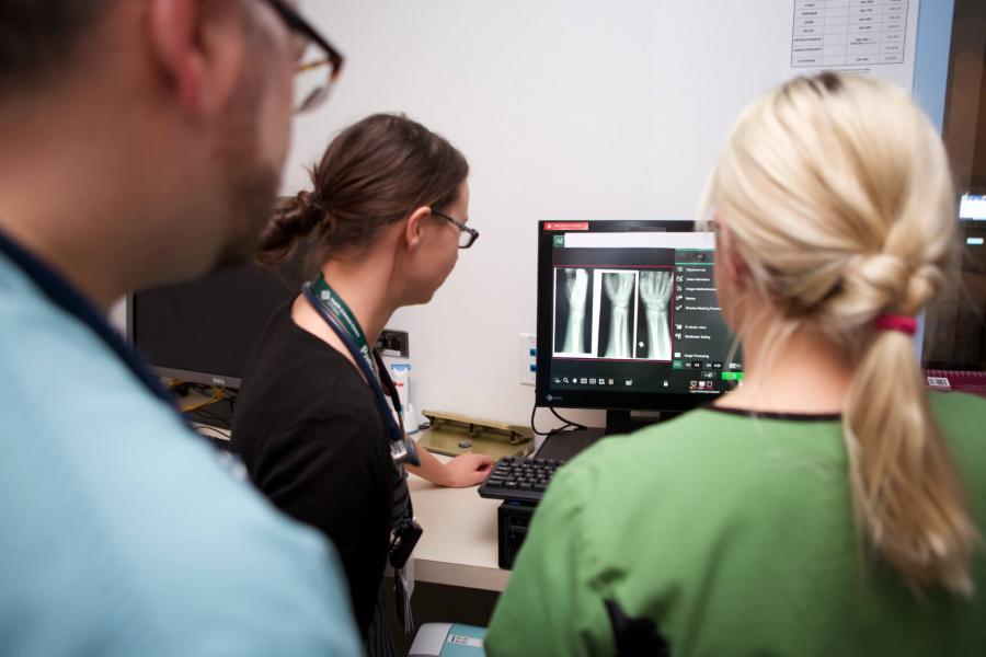 Medical staff gather around a monitor displaying a black and white image of an arm and wrist.