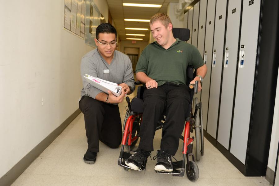 An occupational therapist kneels down in a hallway lined with lockers to show something to a patient in a wheelchair.