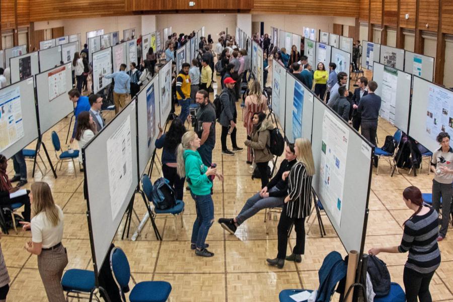 A room filled with undergraduate students standing beside their research posters while a crowd of attendees moves through the room and views everyones work. 
