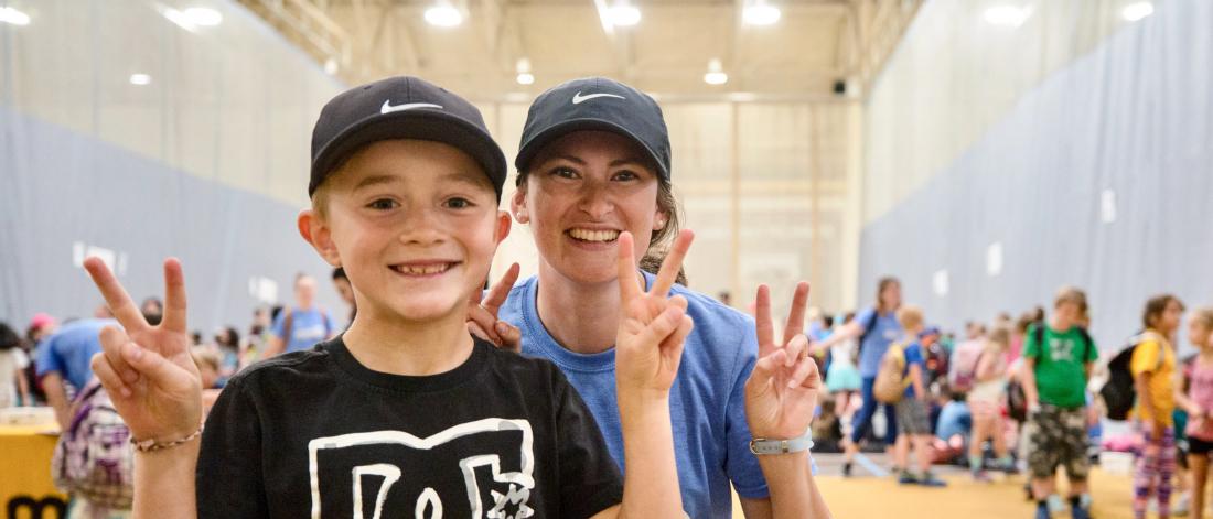 A Mini U leader and child smile at the camera making peace signs in a busy gymnasium.