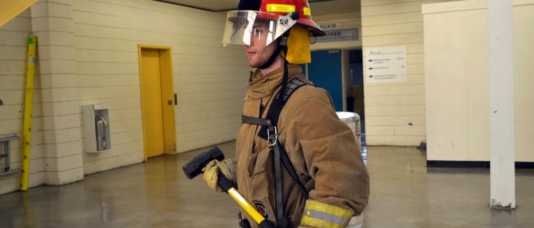 A fireman walks across a room in full gear carrying an axe.