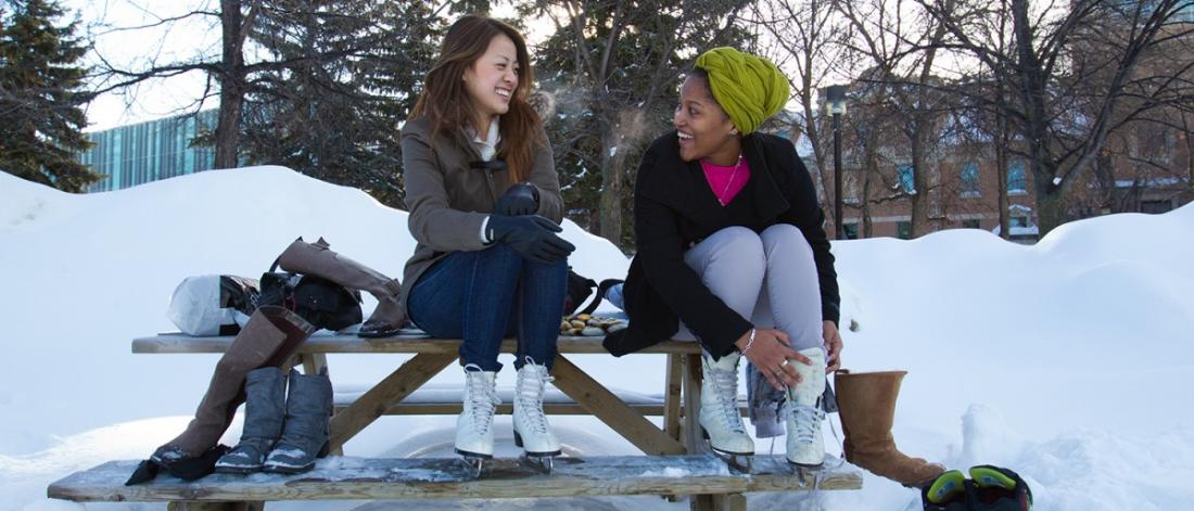 Two young alumni lacing up ice skates on a bench