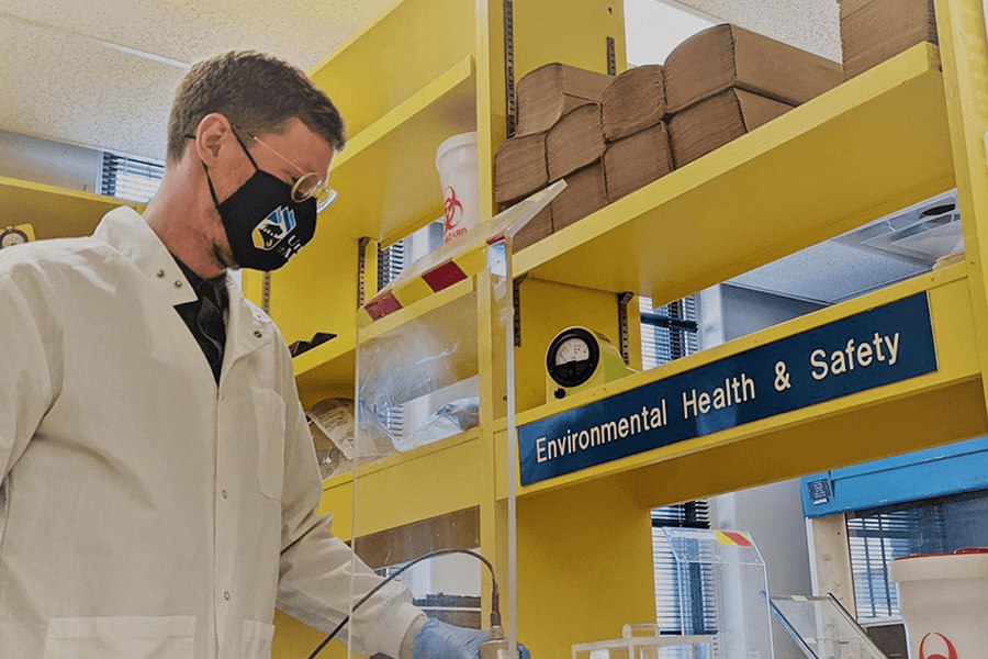 A person in a lab coat working in a lab. A sign on a yellow shelf says "Environmental Health and Safety".