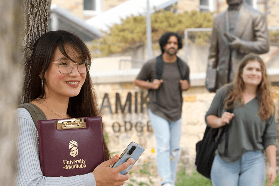 UM student on the foreground holding a burgundy UM branded clipboard. Two other students walking on the background who are out of focus.
