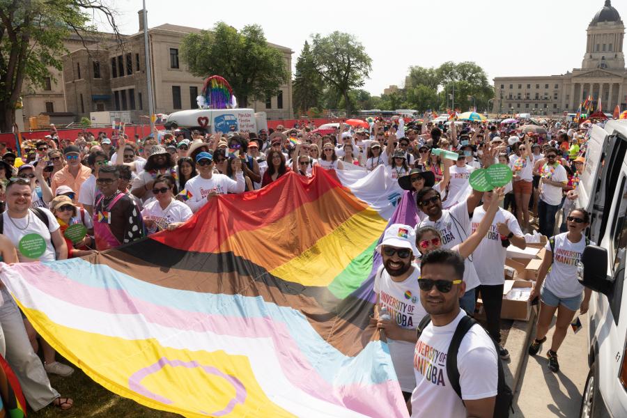tens of people pictured outdoors on a sunny day holding the progressive pride flag