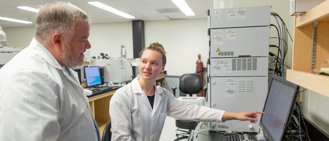 A student points to a computer monitor while talking with a professor in a research lab.