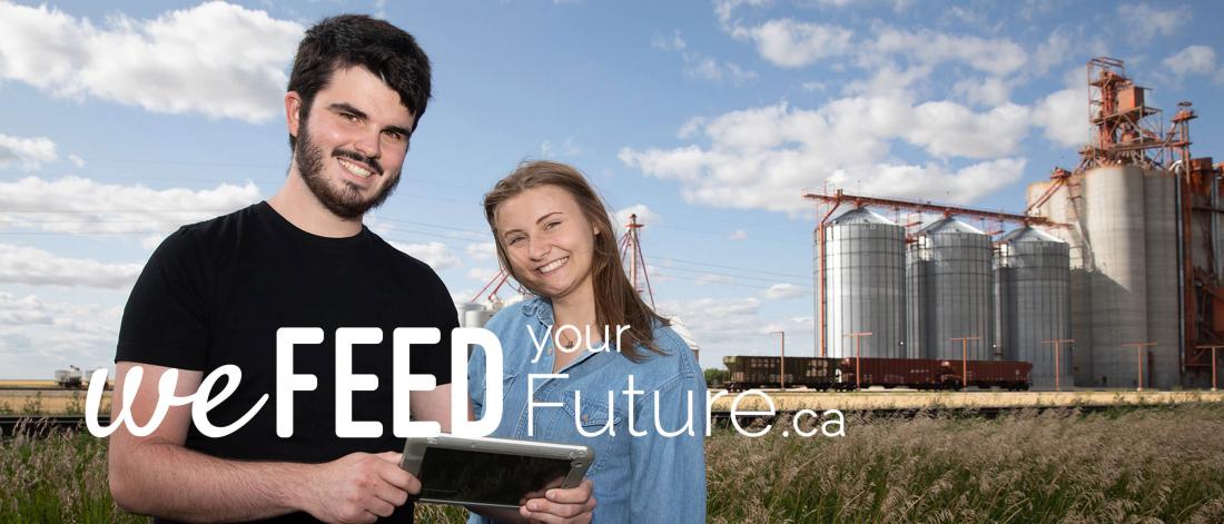 Two students stand near a grain elevator, one holding a tablet.