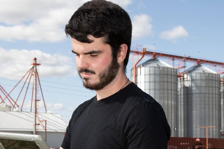 A student uses a piece of equipment while standing in a field with a large grain elevator in the background.