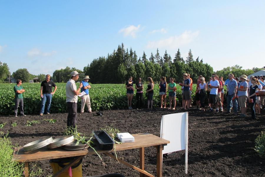 A group of people listen to a researcher standing in a field.