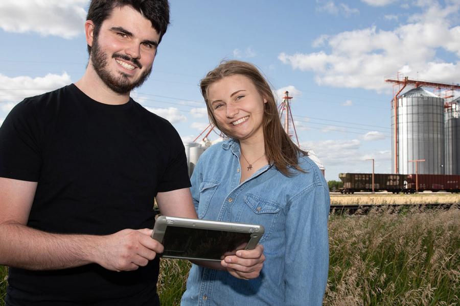 Two students stand near a grain elevator, one holding a tablet.