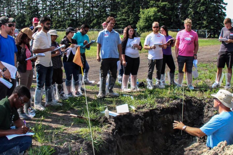 Students gather around an instructor in a hole explaining the soil