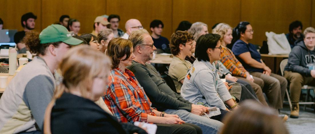 Audience attentively listening during a presentation in a conference room. Attendees are diverse, casually dressed, creating a focused atmosphere.