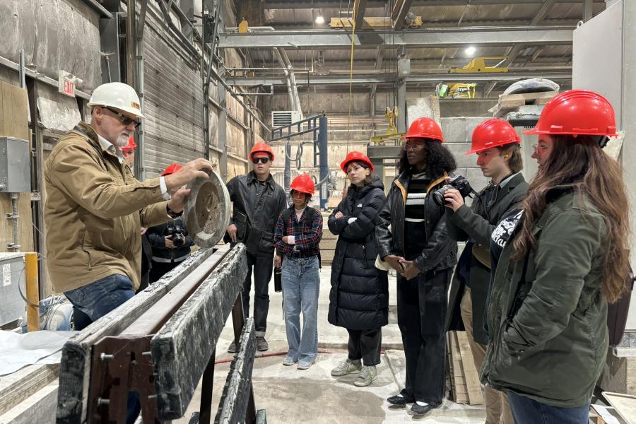 A group of individuals wearing red hard hats watches a man demonstrating a technique while holding a circular object in a manufacturing facility. The setting includes industrial equipment and a partially visible workshop environment.