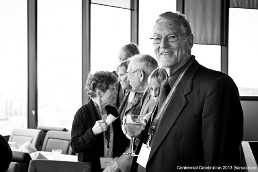 A smiling elderly man in a suit holds a drink, with a group conversing in an elegant, well-lit setting.