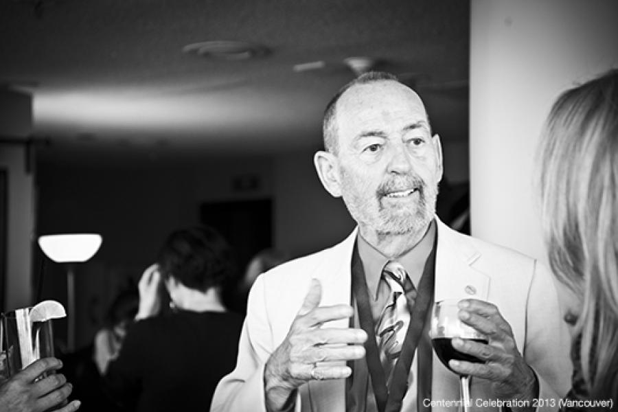 A bearded man in a suit gestures while holding a glass, engaged in conversation in a softly lit room.