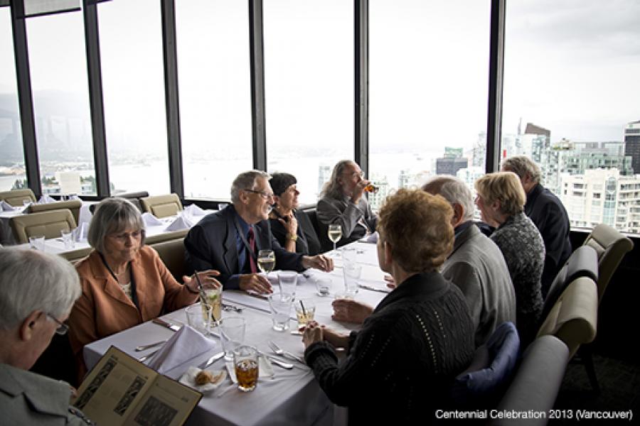 Nine elderly individuals dine around a table, conversing and relaxing in a dimly lit restaurant with panoramic city views.