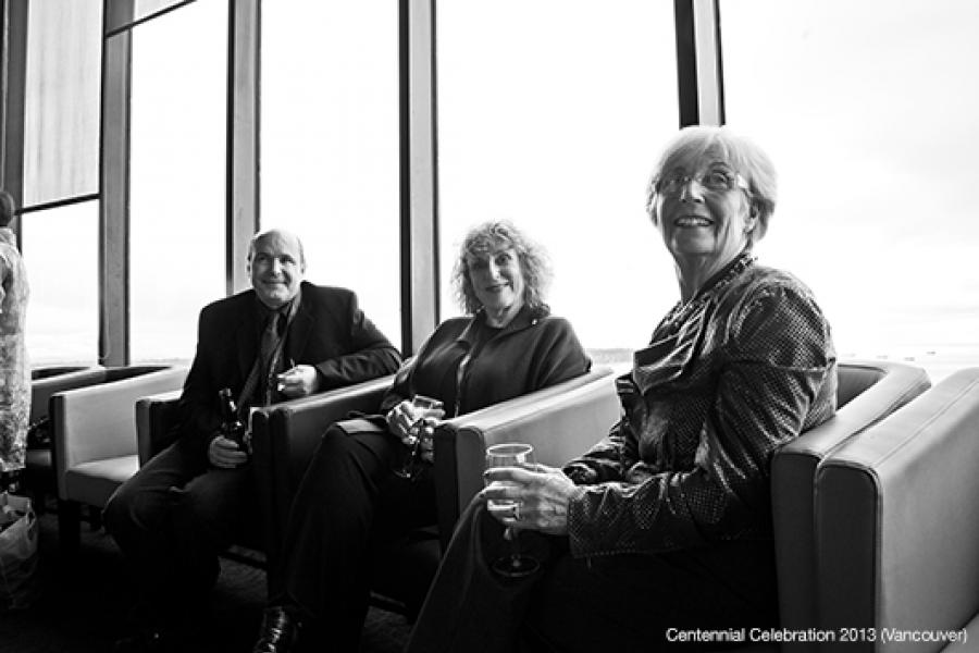 Three individuals in formal attire sit casually with drinks in a modern, well-lit room, exuding a refined, social ambiance.