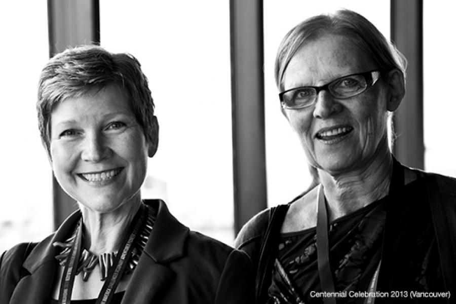Two women with lanyards sit indoors by large windows. One wears a blazer and necklace, the other glasses and a patterned top.