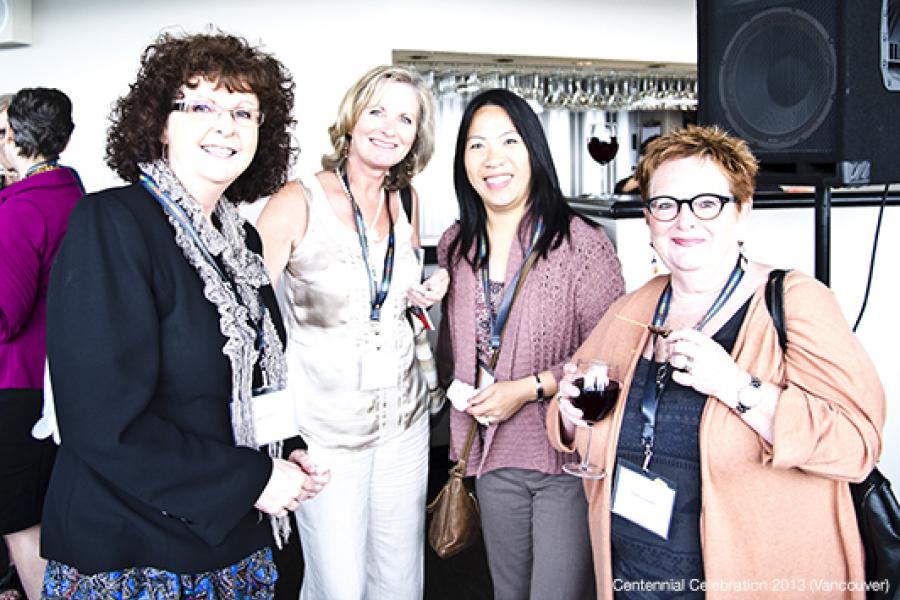 Four women at a bright networking event, dressed stylishly, with drinks in hand, chatting in a convivial atmosphere.