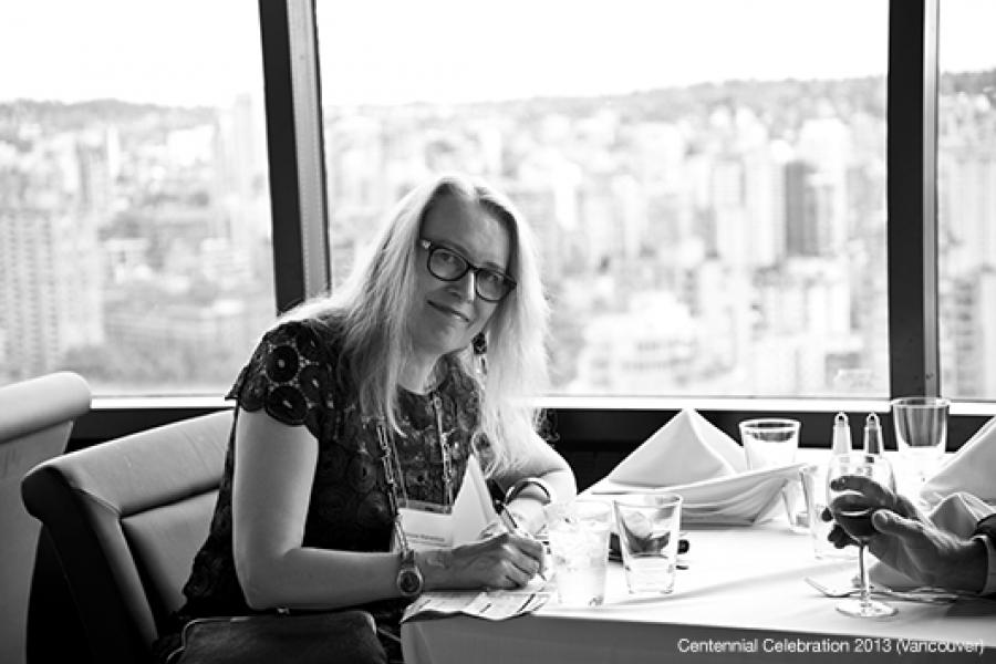 A woman with long hair and glasses writes at a restaurant table, overlooking a cityscape through a large window.