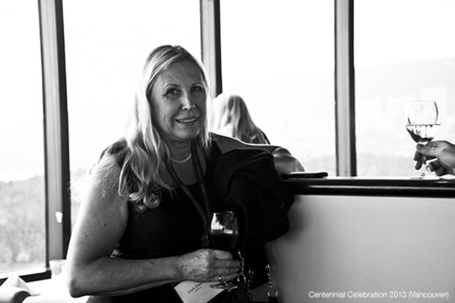 A smiling woman with long blonde hair holds a glass of wine, standing by a railing in a bright, well-lit room.