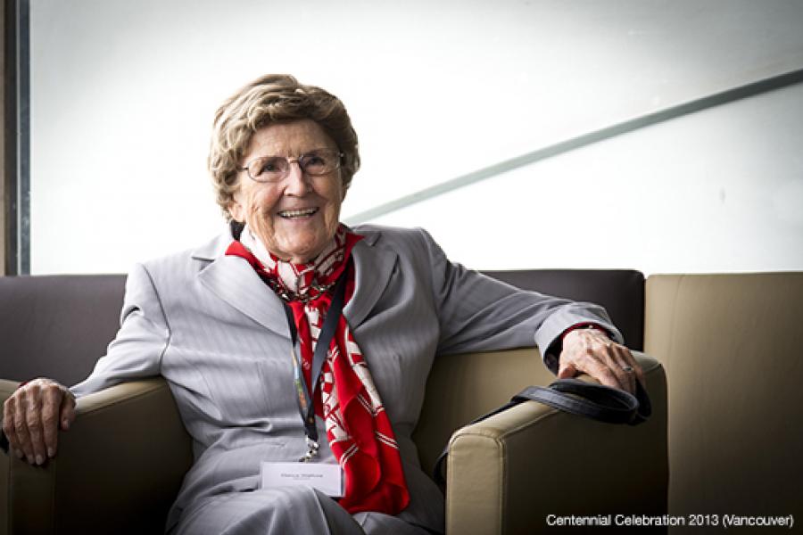 An elderly person in a light gray suit and red scarf smiles warmly while seated in a modern armchair in a minimalist setting.