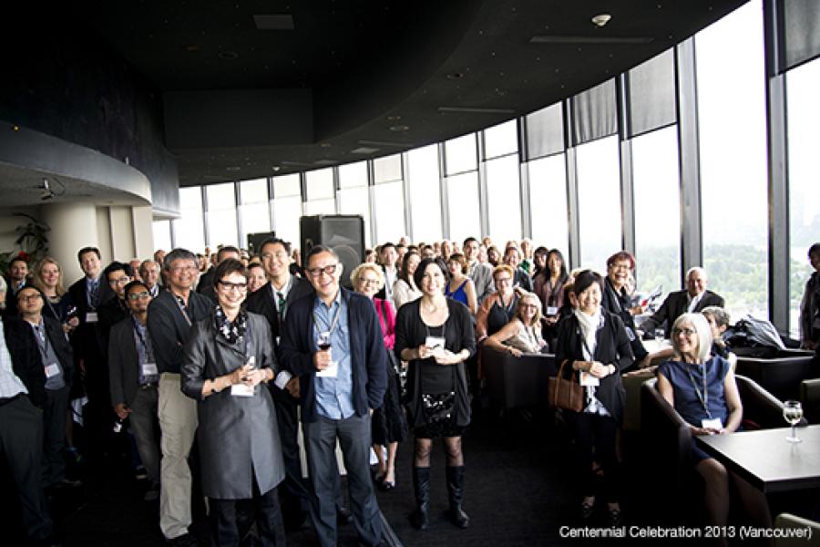 A diverse group of people in business attire gathers in a bright, modern high-rise with expansive windows, suggesting a social event.