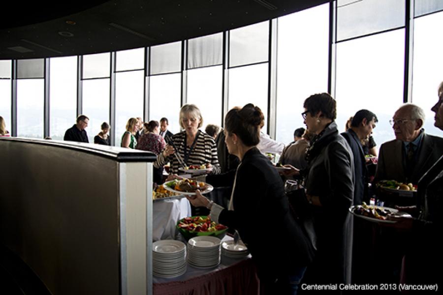 People in formal attire gather in a spacious room with large windows for a buffet-style event, interacting and serving themselves food.