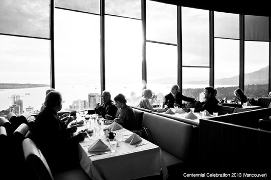 In a sophisticated high-rise restaurant, diners sit at elegantly set tables, enjoying meals while overlooking a panoramic city and coastline view.