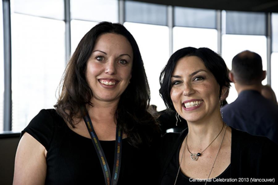 Two smiling women in black attire stand closely together indoors, illuminated by natural light from large windows, creating a cheerful atmosphere.