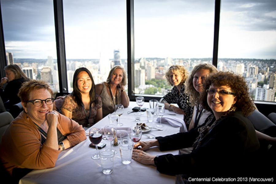 Six cheerful women sit around a white tablecloth-covered table in a restaurant, enjoying conversation with a panoramic city skyline view outside.