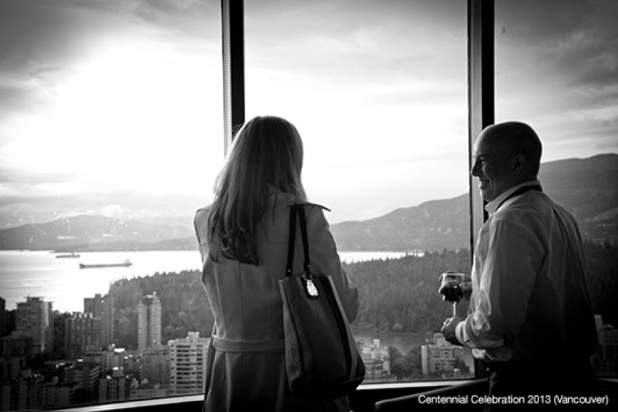 A person with long hair in a coat faces away, holding a handbag, while another holds a wine glass, engaged in conversation by a window overlooking a cityscape.