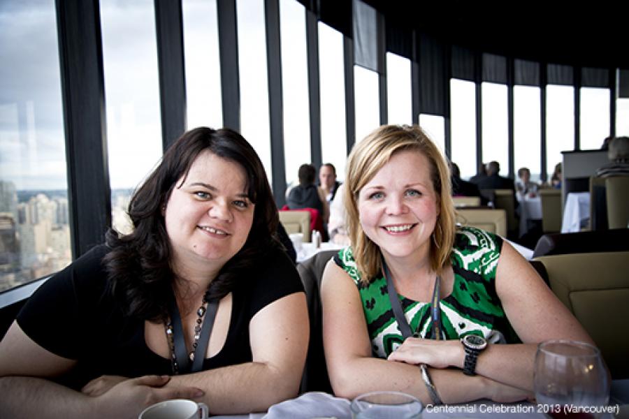 Two women sit at a table in a bright restaurant, smiling and leaning forward. The large windows behind them offer a panoramic cityscape view.