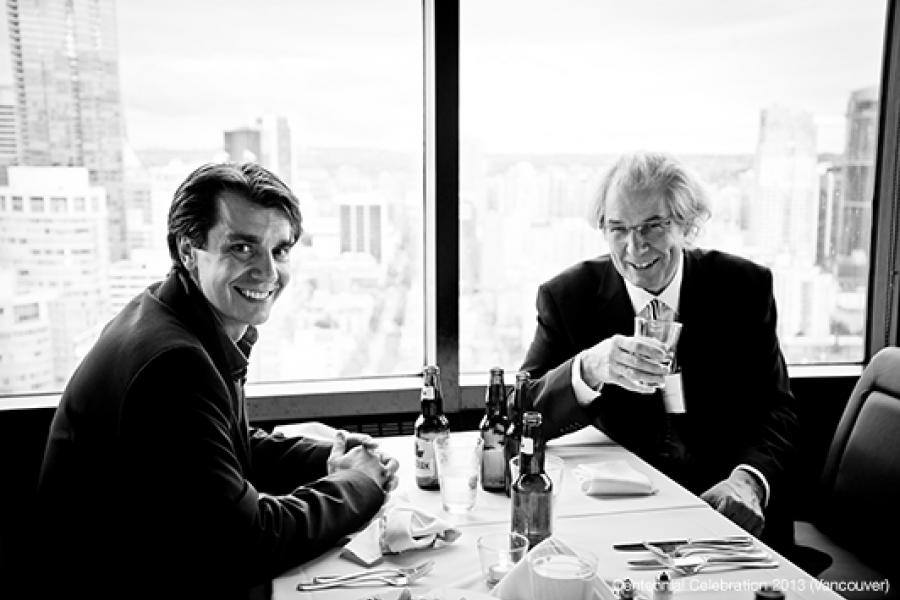 Two men sit at a table in a high-rise restaurant, smiling and gesturing as if toasting, with a cityscape view in the background.