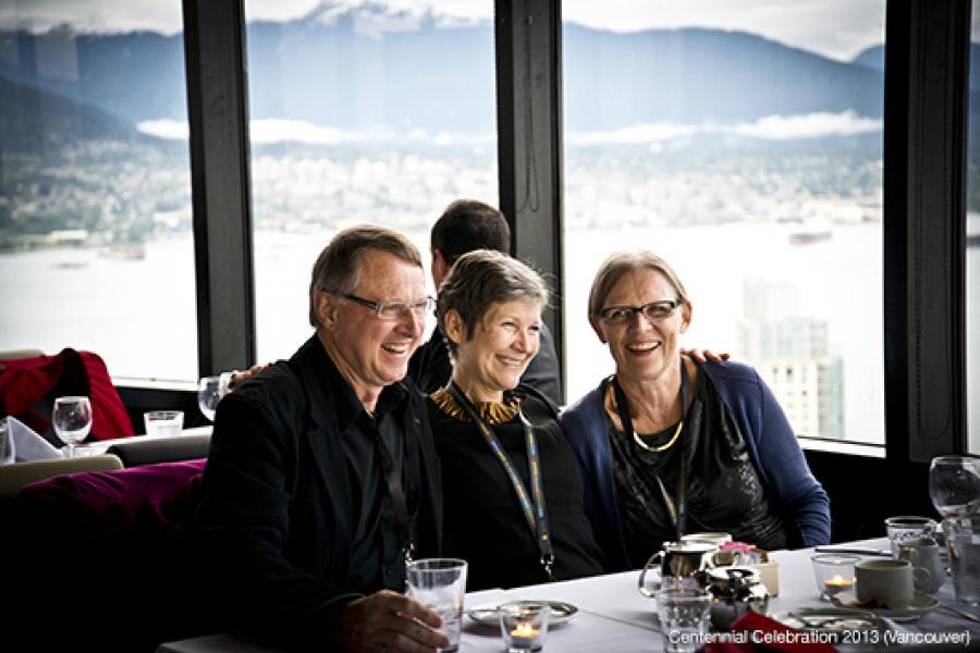 Three smiling individuals sit closely at an elegantly set dining table, enjoying a social occasion with a scenic city and mountain view behind them.