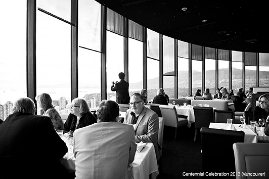 A sophisticated high-rise restaurant features floor-to-ceiling windows with a cityscape view. Elegantly dressed patrons engage in conversation at white-clothed tables, enjoying their meals in natural light.